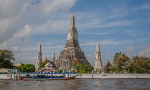 Explore the iconic Wat Arun temple along the Chao Phraya River in Bangkok under a vibrant sky.